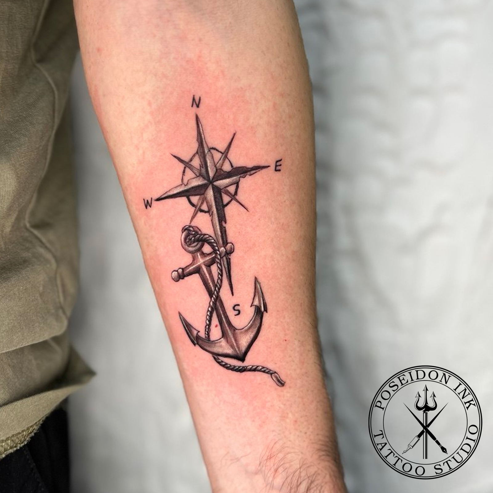 Black and grey realism tattoo of an anchor wrapped in rope over a labelled compass rose, on the forearm