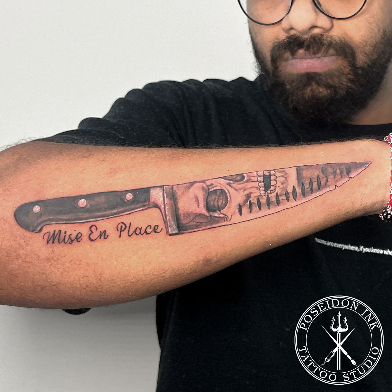 Realistic black and grey tattoo of a chef's knife with a screaming skull reflected in the blade and Mise En Place script