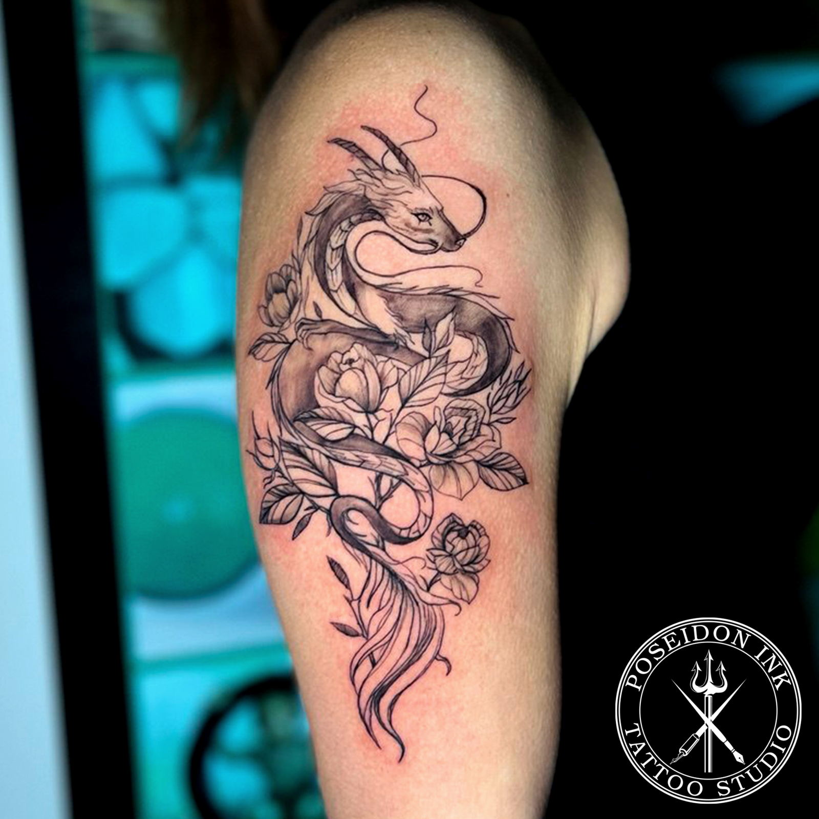 Fine line tattoo of a serpentine dragon with flowing mane coiling through peony flowers on the upper arm