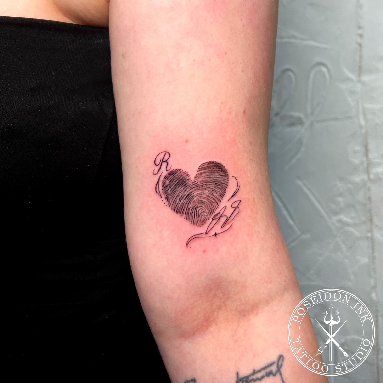 Fine line tattoo of a heart made from two overlapping fingerprints with script initials on the upper arm