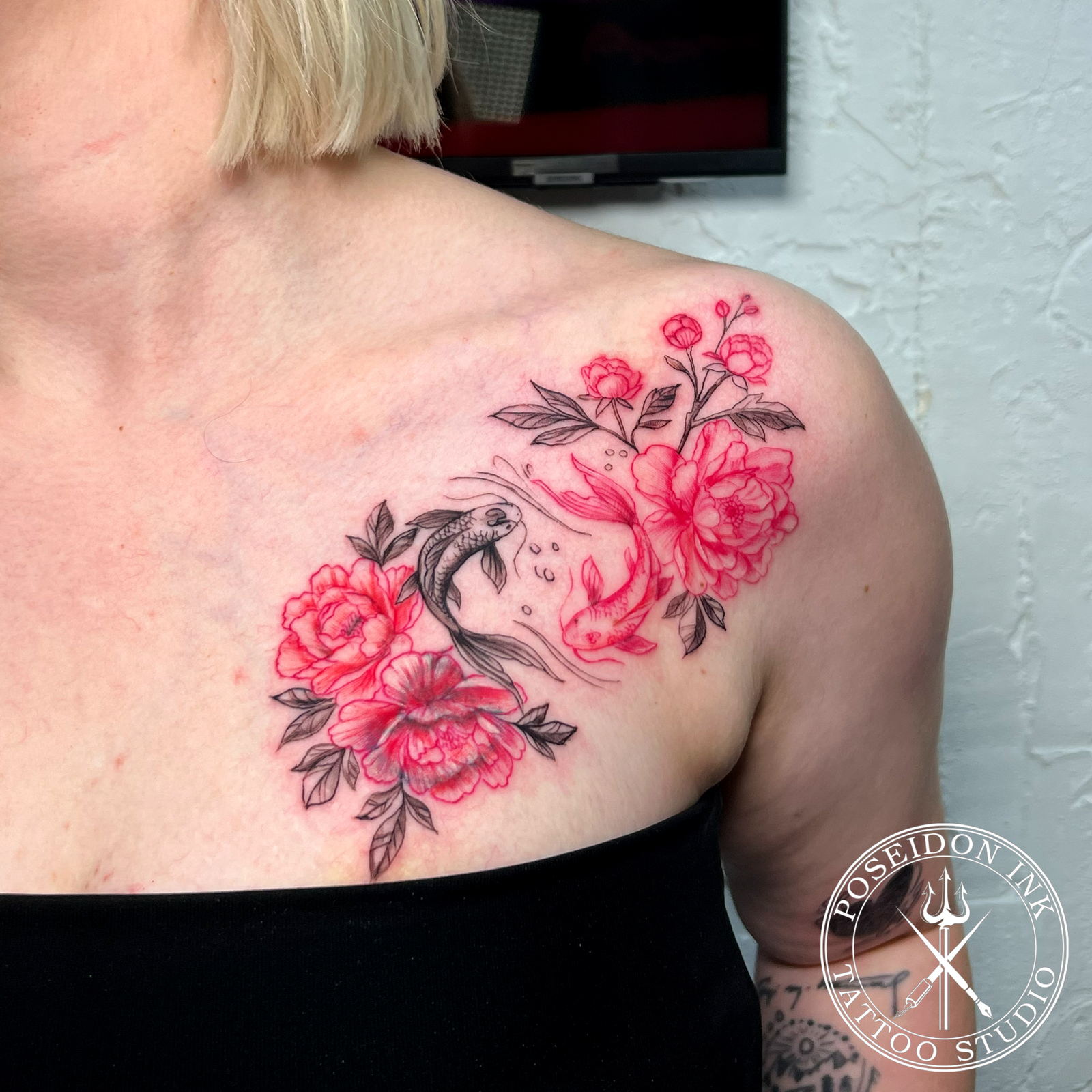 Fine line tattoo of a black koi and red koi circling among red peonies, flowing across the collarbone and shoulder