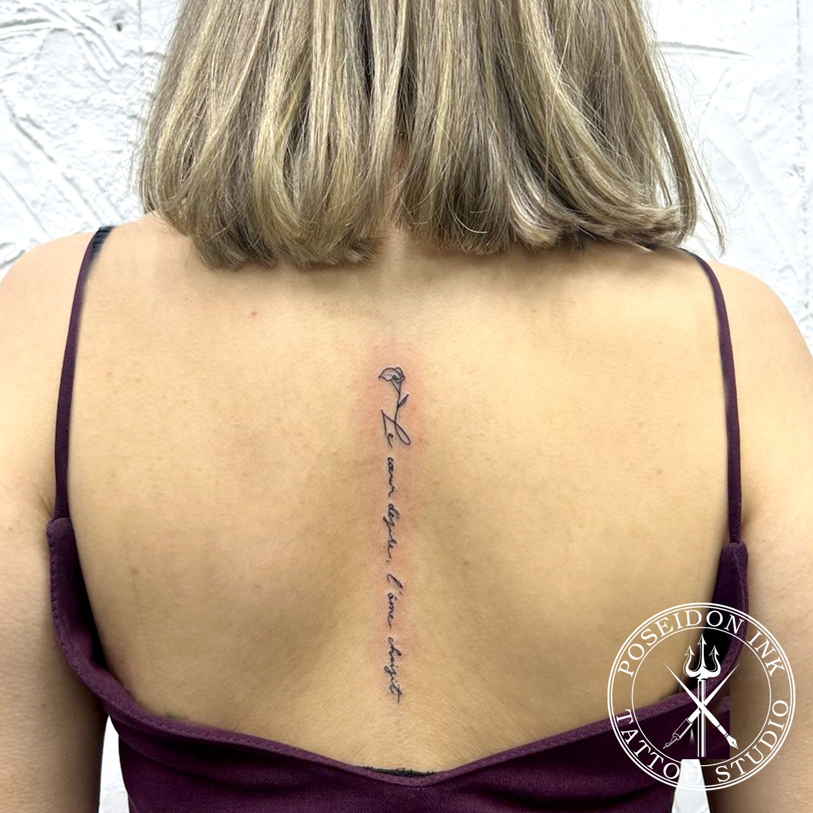 Fine cursive lettering tattoo running down the spine, topped with a small fine-line lily flower