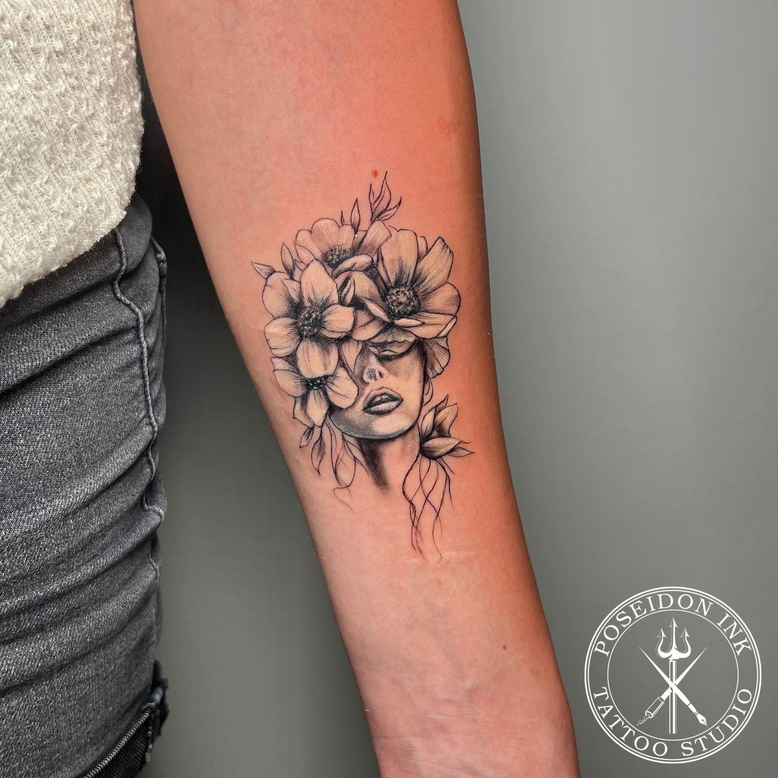 Surreal black and grey tattoo of a woman's face with anemone flowers replacing the upper half of her head, forearm