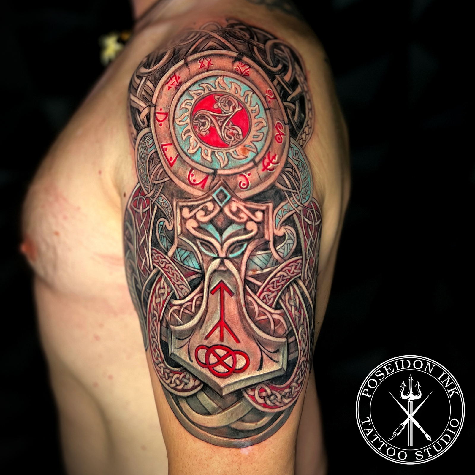 Colour tattoo of Thor's hammer with Celtic knotwork, red runes and a triskelion sun disc in carved 3D stone effect, upper arm