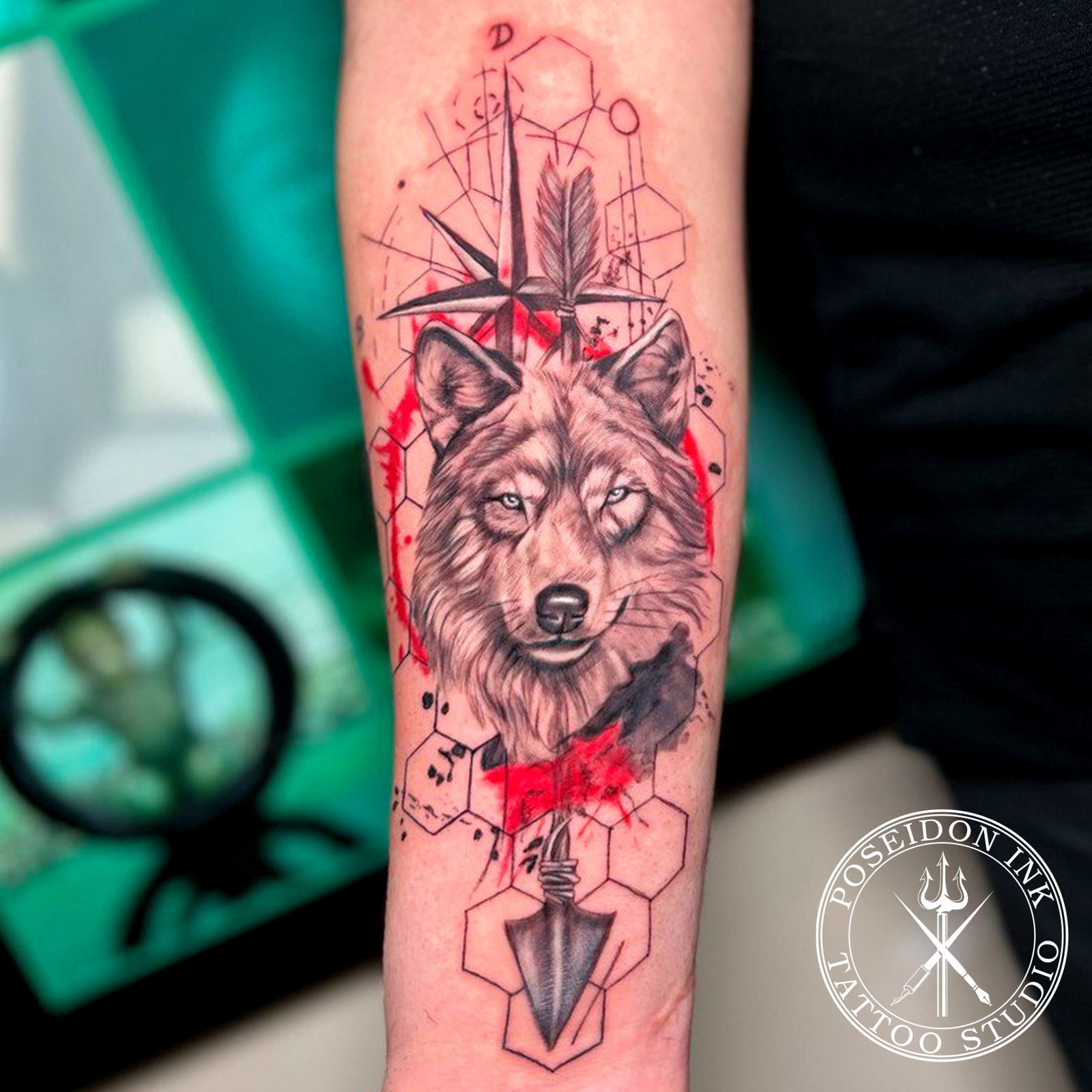 Black and grey realistic wolf tattoo with compass star, feathered arrow, hexagons and red trash polka strokes on the forearm